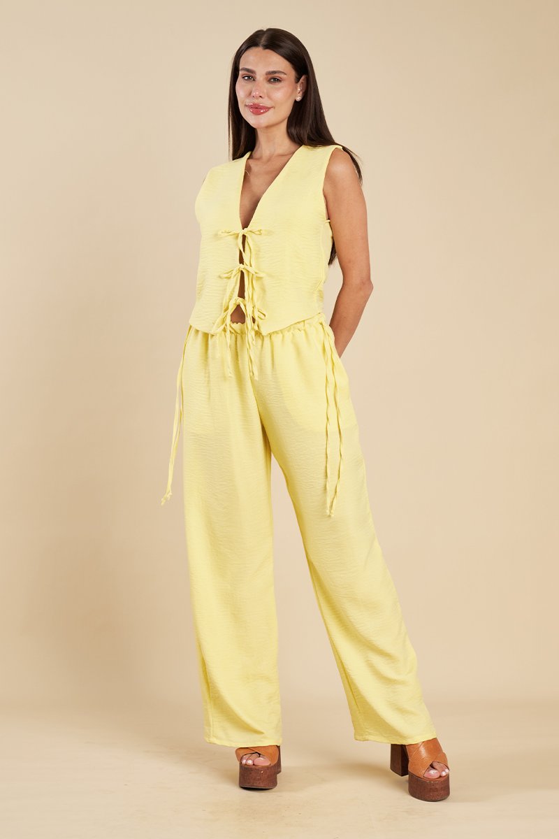 Detalhe de calça amarela de perna larga com cordão lateral solto, combinada com colete amarelo e bolsa de palha