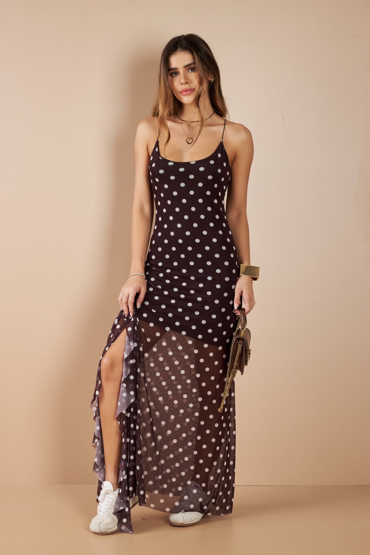 Mulher de vestido longo marrom de tule com poás brancos e alças finas, segurando bolsa marrom de corrente.