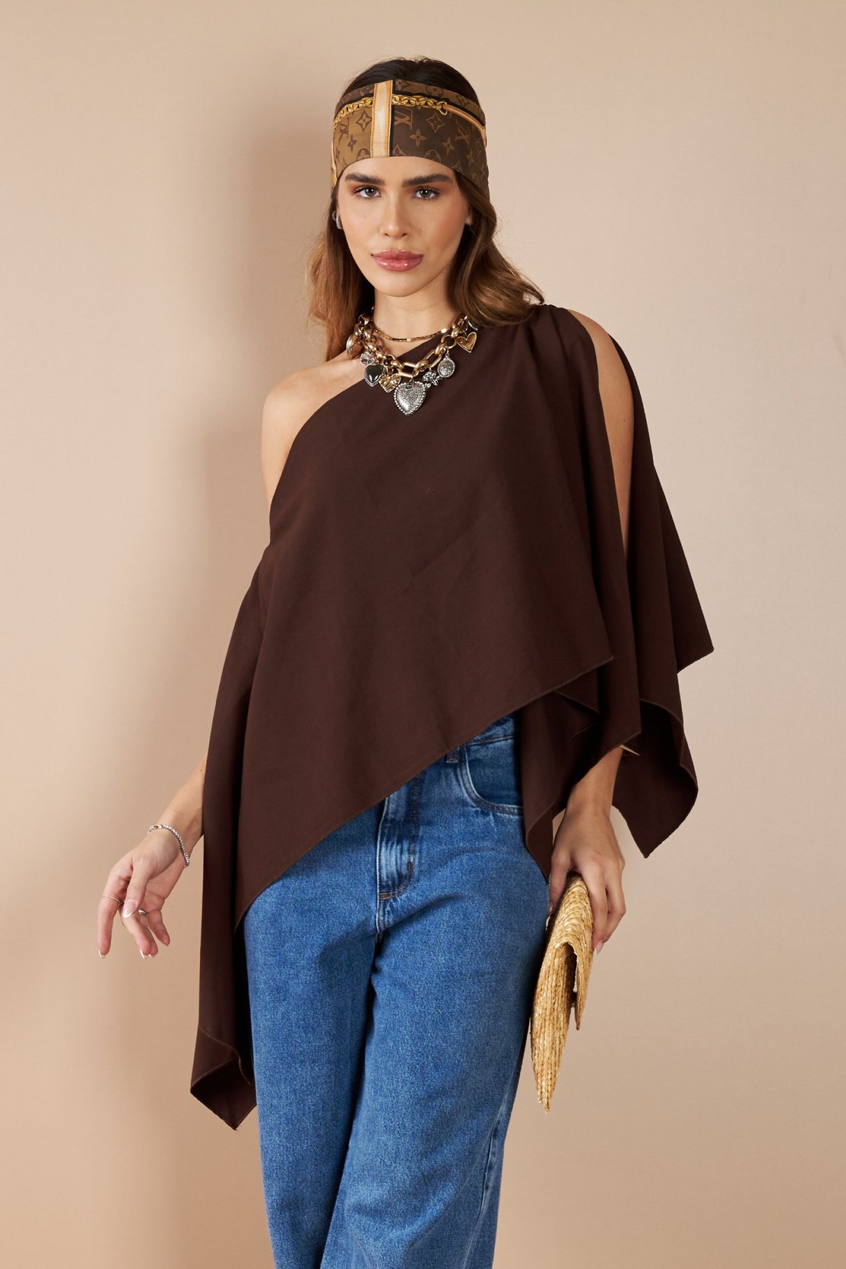 Modelo de poncho marrom de alfaiataria com um ombro descoberto, calça jeans e clutch de palha.
