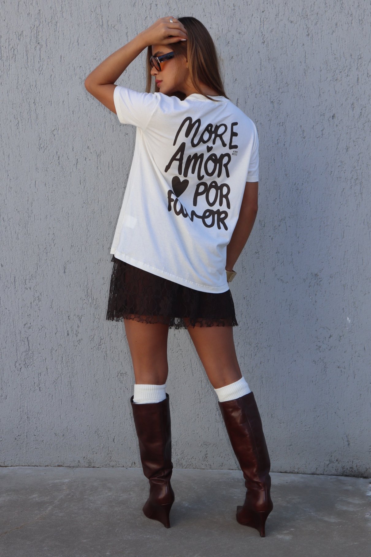 Modelo de camiseta off-white e óculos escuros usa a saia curta marrom de renda com meia branca e bota de cano alto