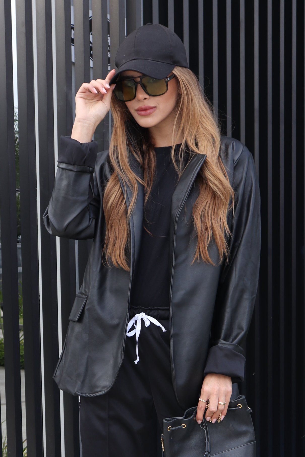 Modelo de boné e óculos escuros usa blazer preto de couro rayon aberto sobre camiseta e calça pretas, diante de painel ripado escuro